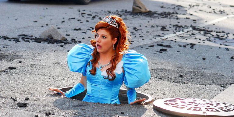Nicole Parker, dressed in a blue princess dress, pops up from a manhole cover in the road.