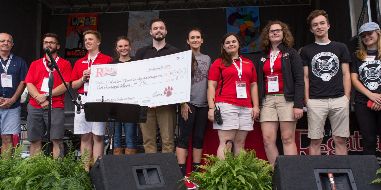 A group of people stand on a stage during a presentation with a giant scholarship check.