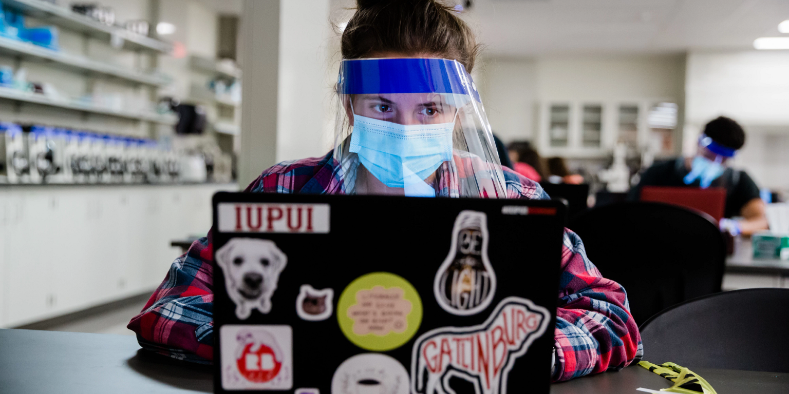 a student wearing a face mask and shield works on their computer