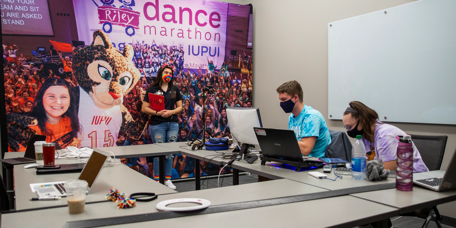 Two students sit around table with laptops and one student stands in front of a Jagathon banner.