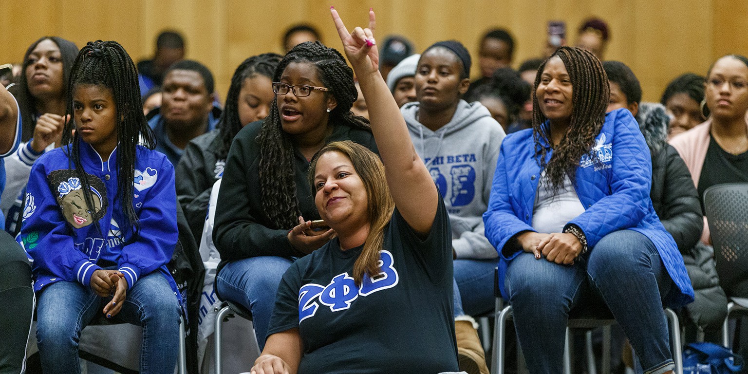 A woman sits in the crowd holding up the hand sign for her sorority, Zeta Phi Beta Sorority