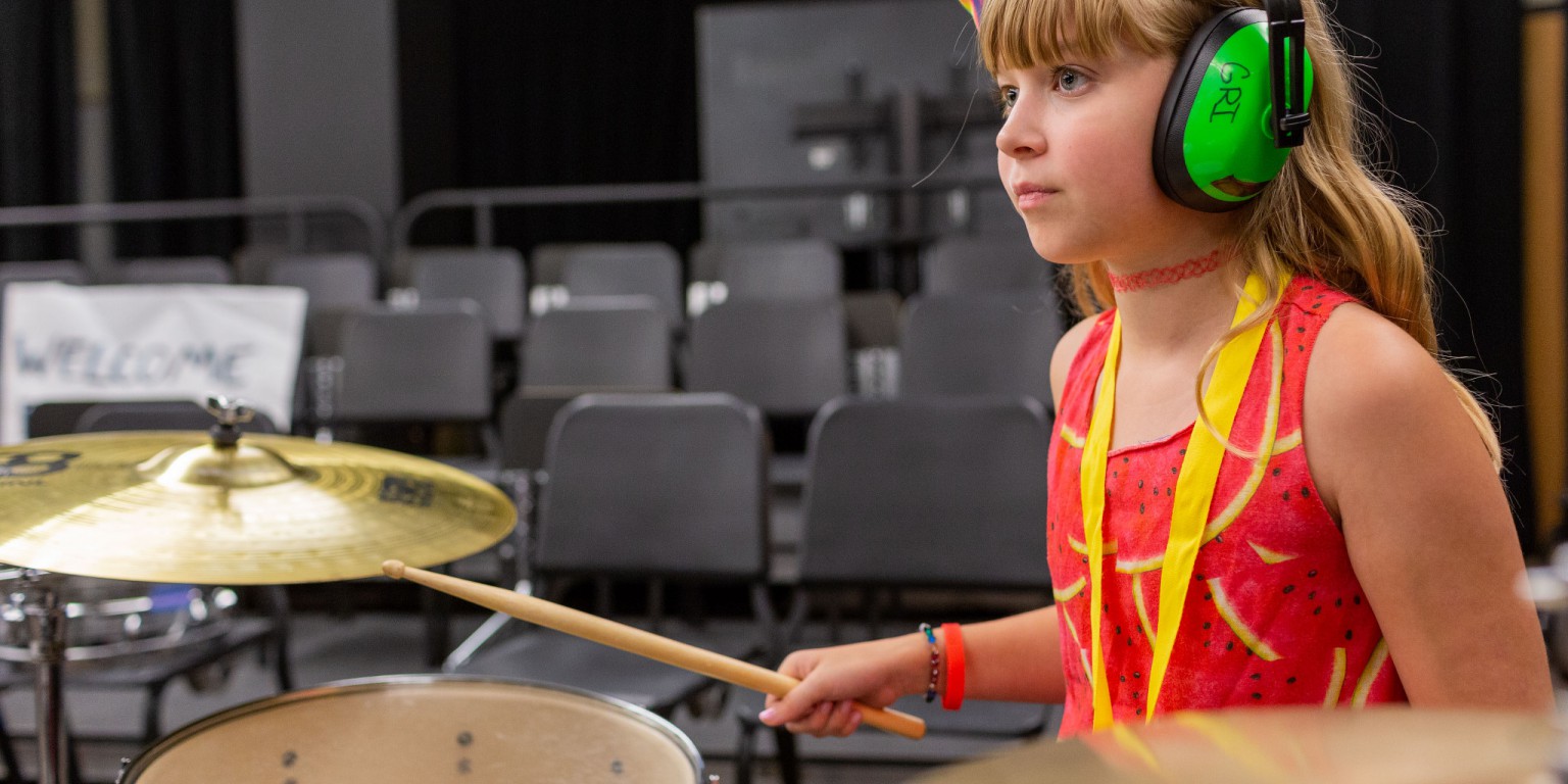 A Girls Rock Indianapolis camper in an orange-and-yellow tank top rocks the drums.