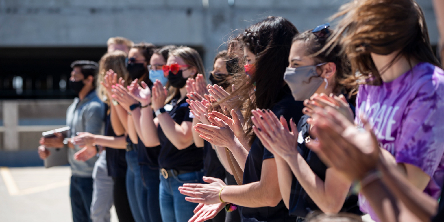 Students wearing masks stand in a line clapping.