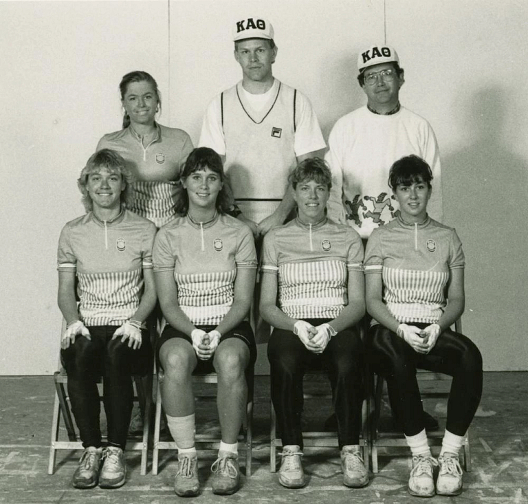 Four women in cycling uniforms sit in chairs while another woman and two men stand behind them.