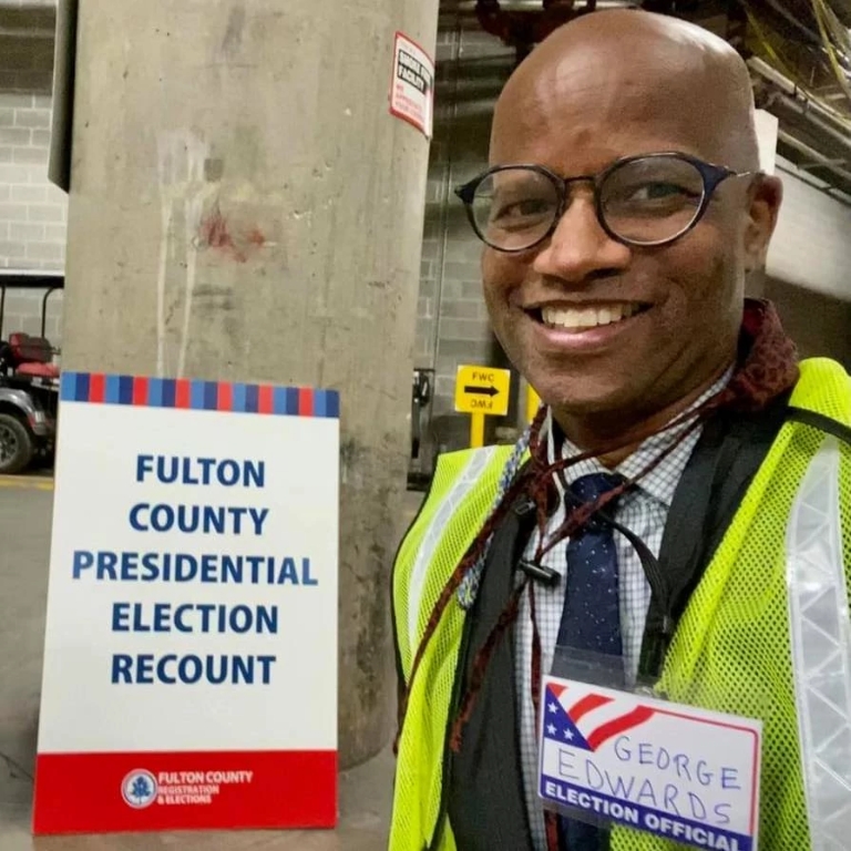 a man stands in front of a sign that says Fulton County Presidential Election Recount