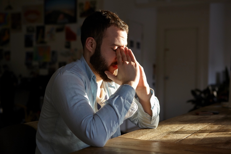 A man sits alone at a table in a darkened room with his face in his hands.