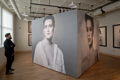 A man in a suit stands and looks at a mural-sized photograph of an androgynous model.