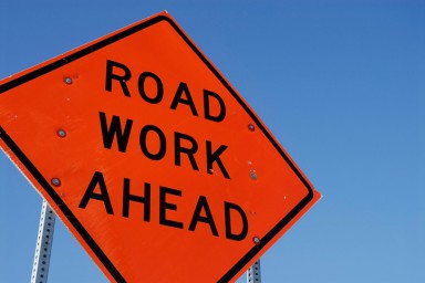 An orange road work sign against a blue sky.