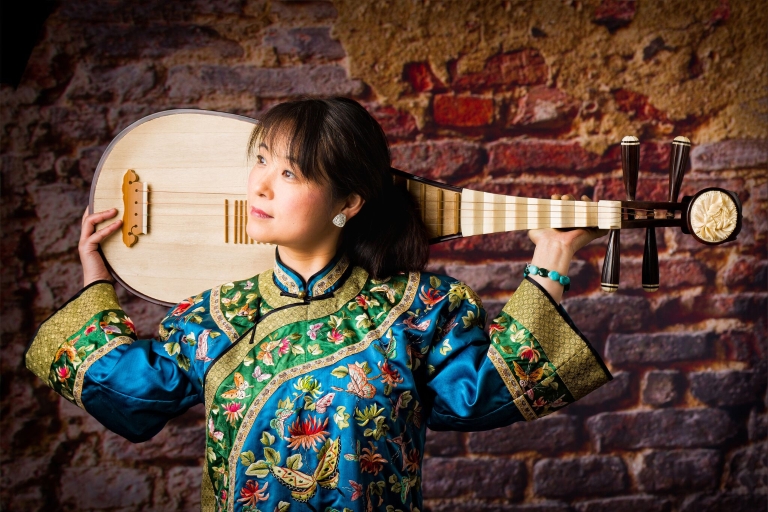 Musician Wu Man posing with her instrument, a pipa, across her shoulders in front of a brick wall.
