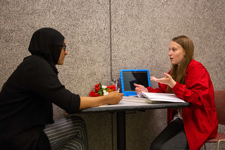 A student sits at a table helping another student with her speech