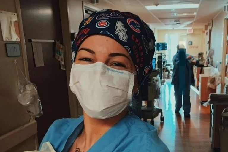 Nurse smiles in hospital hallway wearing scrubs, a Cubs head covering and a blue mask