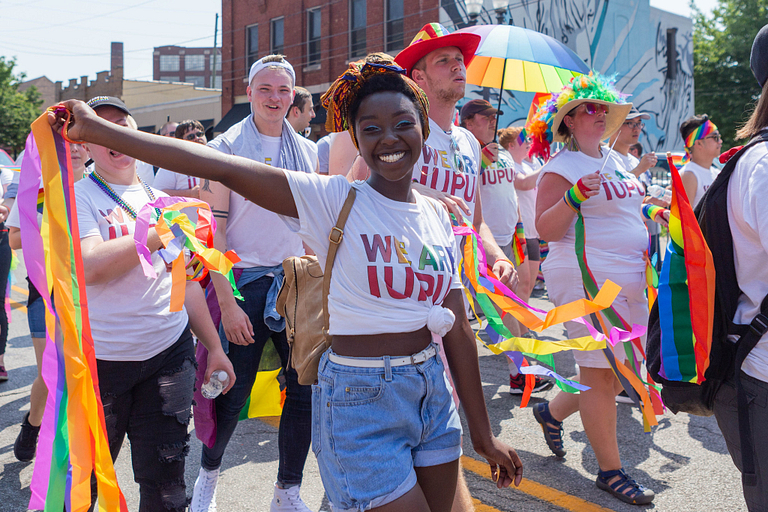 A female IUPUI student wears an 'I am IUPUI' shirt and dances with rainbow streamers.