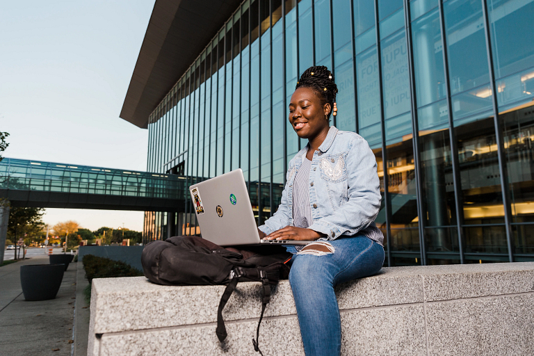 A female student looks at her laptop while sitting outside a building