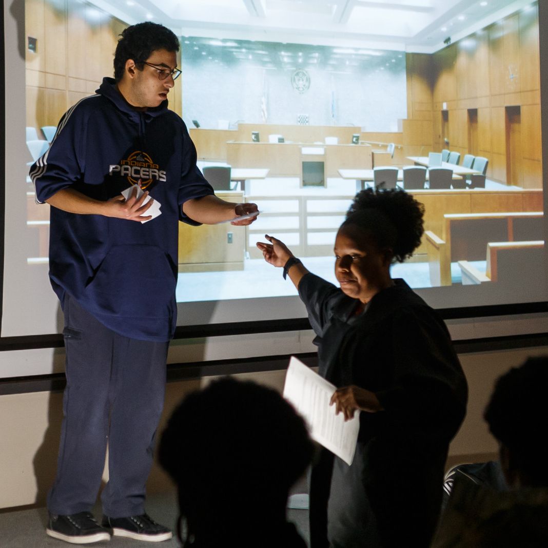 One person is on a stage with a courtroom screen behind him, and another person hands him a ticket.