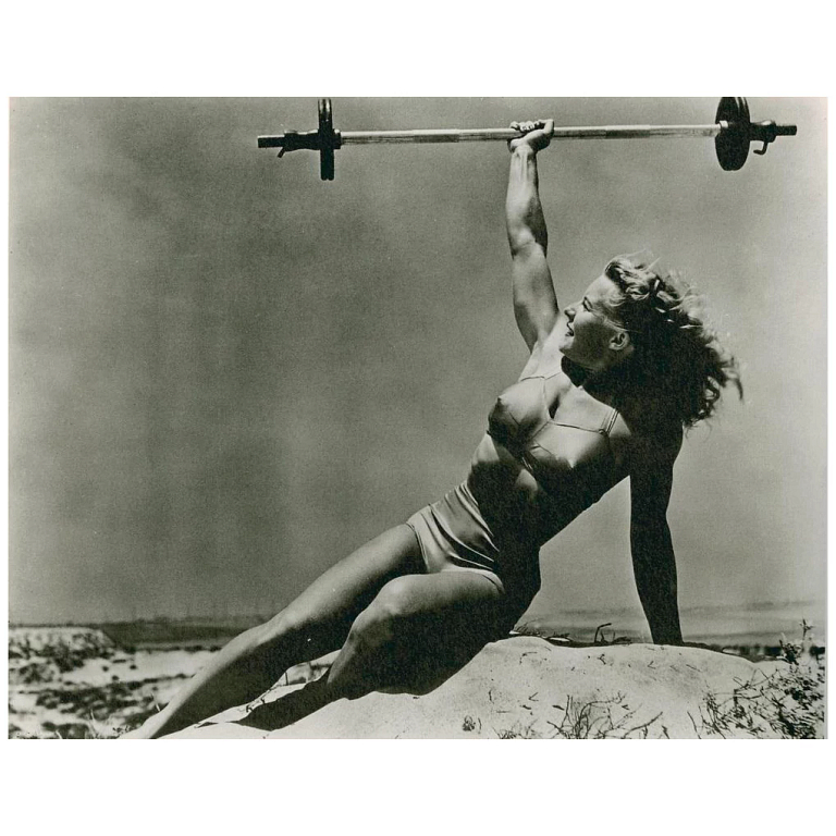 Woman in a bikini sits on a beach lifts a barbell overhead in one hand