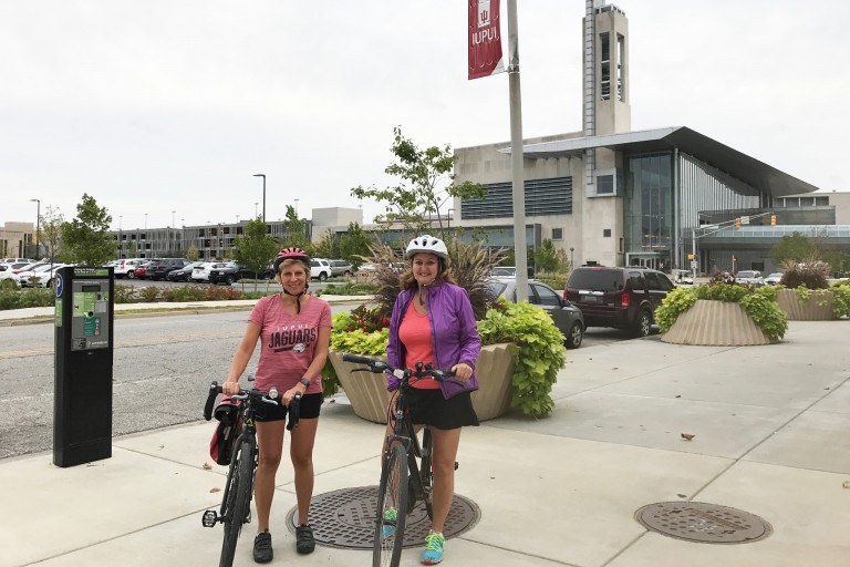 Kathy Johnson and Amy Warner pose with their bicycles with the Campus Center as a backdrop.