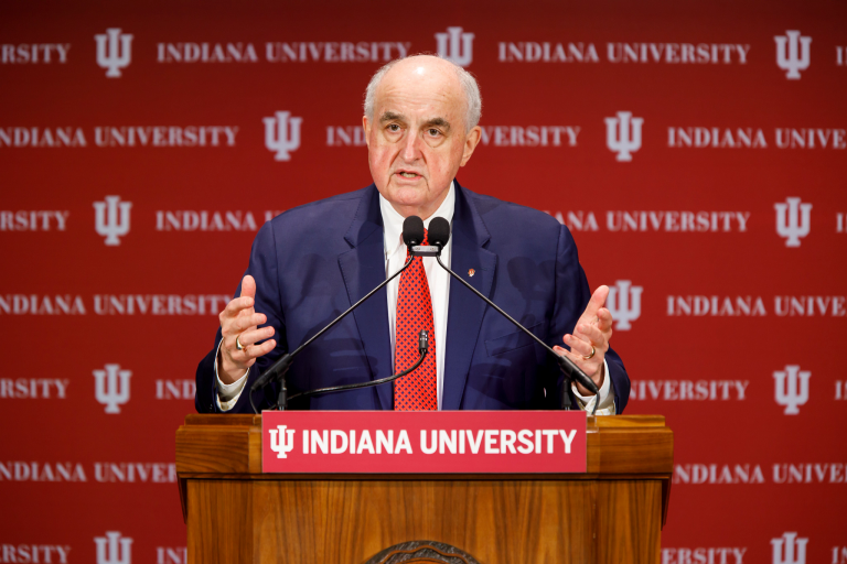 IU President McRobbie speaks at a podium in front of a cream-and-crimson IU background