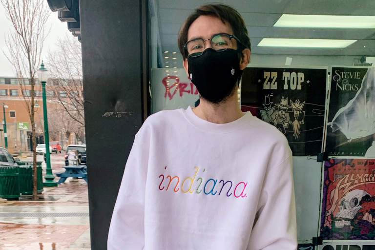Student stands outside Tracks wearing a white sweatshirt with a rainbow "Indiana" embroidered on it
