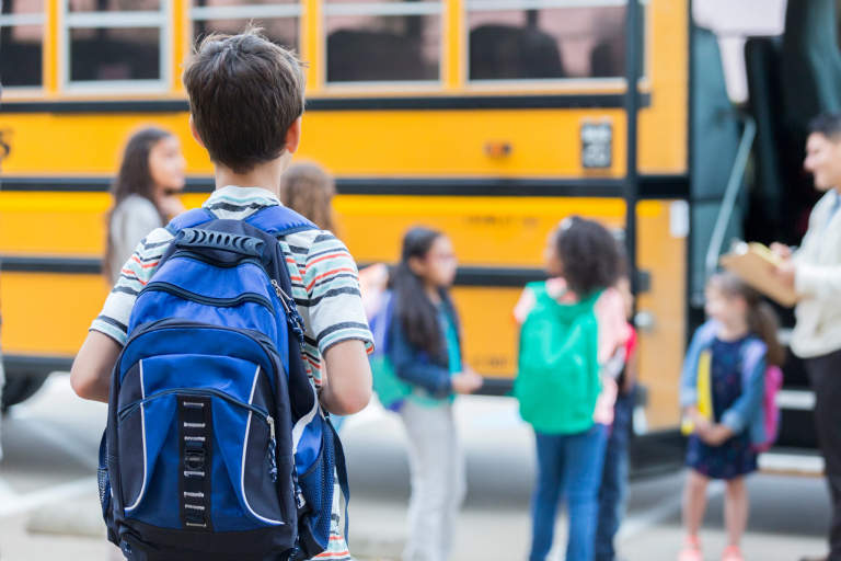 Elementary students wearing backpacks wait to board a school bus