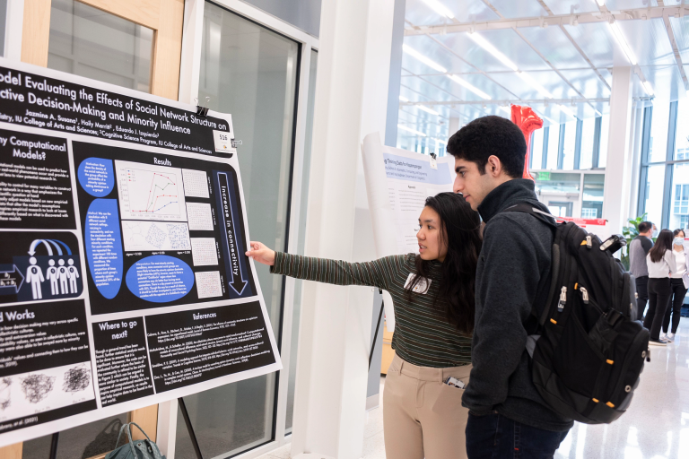 A female student points to her poster while talking to a male student