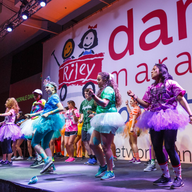 Students in multicolored costumes dance on a stage.