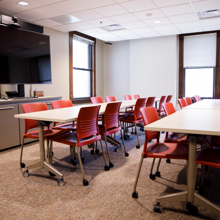 A conference room space, red chairs pushed under two long tables and a large TV on the wall