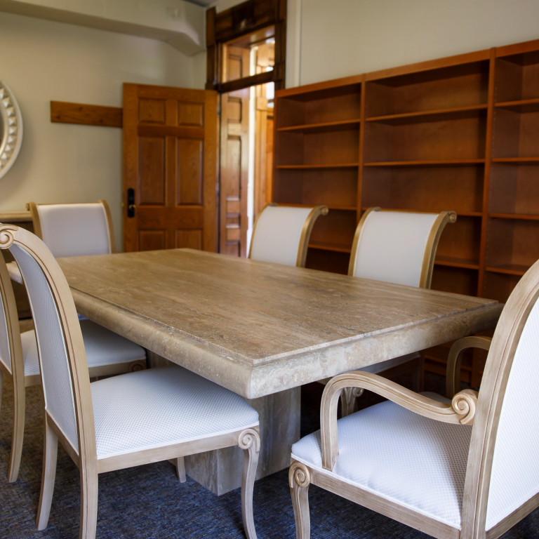 A study with a large marble table surrounded by white chairs, and bookshelves in the background