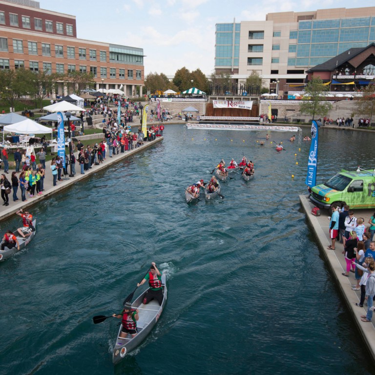 Canoes race on the downtown canal while spectators stand along the water's edge.