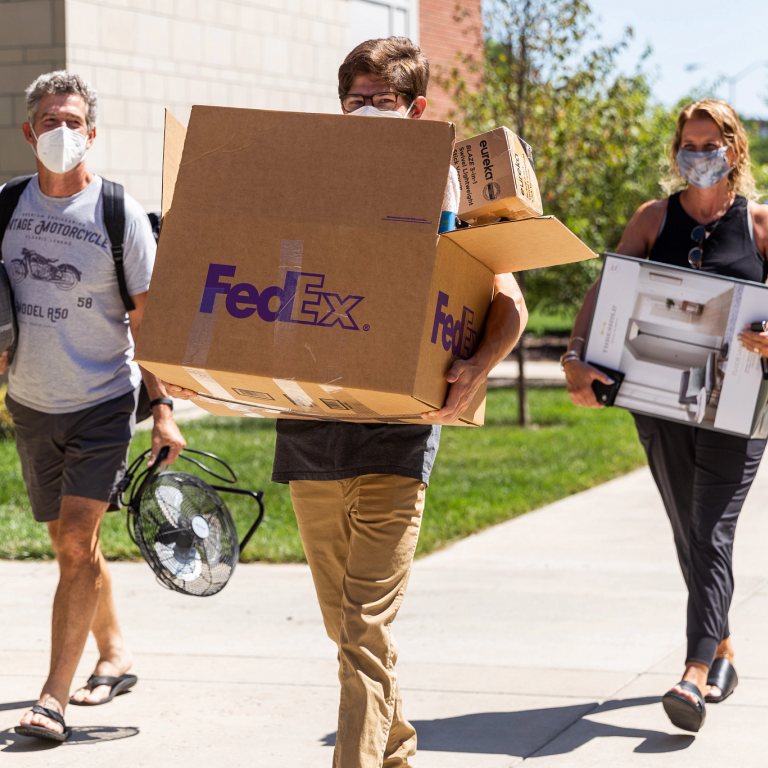 a student and two parents wearing masks carry things in on move-in day