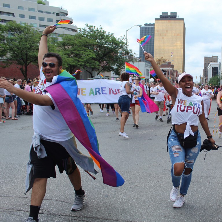 Two IUPUI parade participants smile and wave pride flags during the 2019 Indy Pride parade.