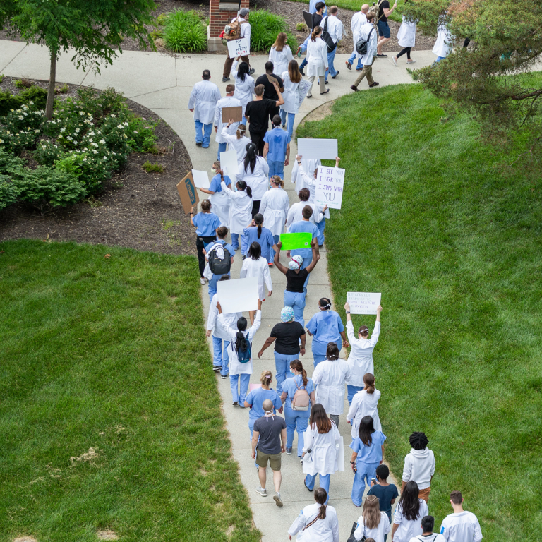 A view from above shows a large group of marchers walking on the sidewalk