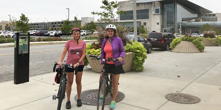 Kathy Johnson and Amy Warner pose with their bicycles with the Campus Center as a backdrop.