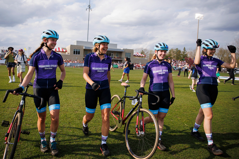 Four women riders in helmets walk through a field in the middle of a cycling track.