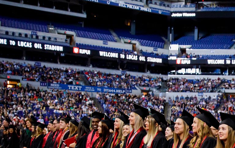 Graduates stand at Lucas Oil Stadium during an IUPUI commencement ceremony.