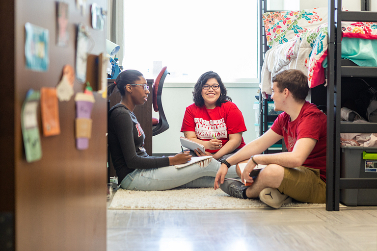 Three students sit on the floor, talking in a dorm room.