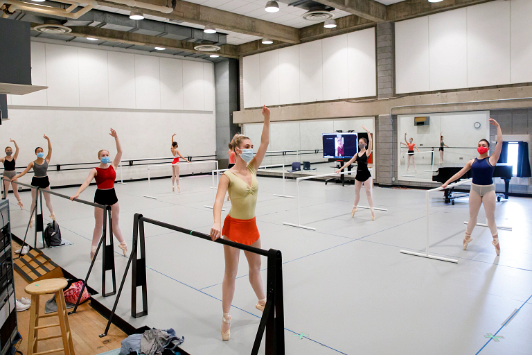 Ballet dancers practice while wearing masks, with the professor on a monitor in the background