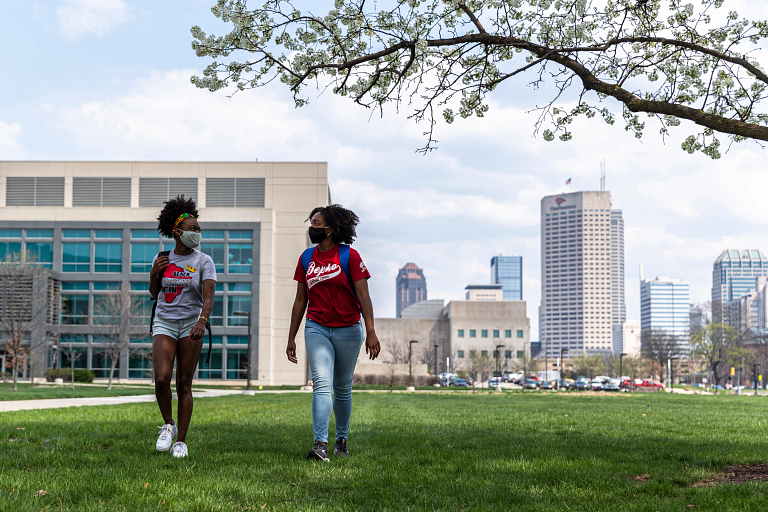 Two students in masks walk on the IUPUI campus, with the Indianapolis skyline in the background