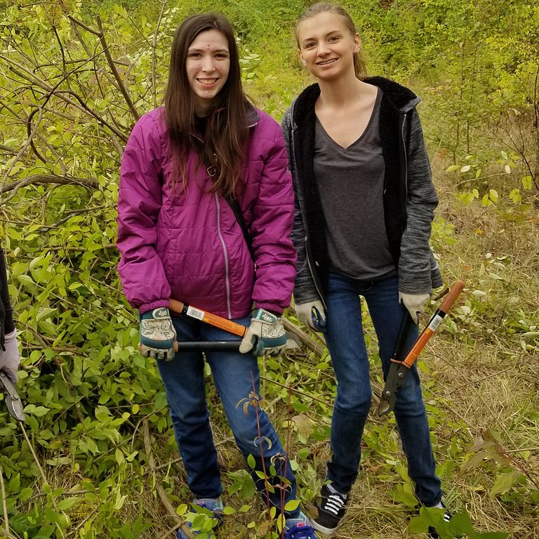 Two female students stand in the woods, preparing to do service work on a trail.