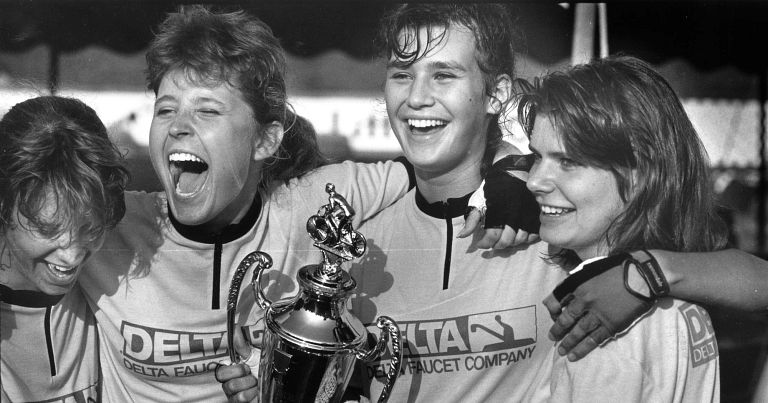 Four women hug each other and celebrate while holding a trophy.