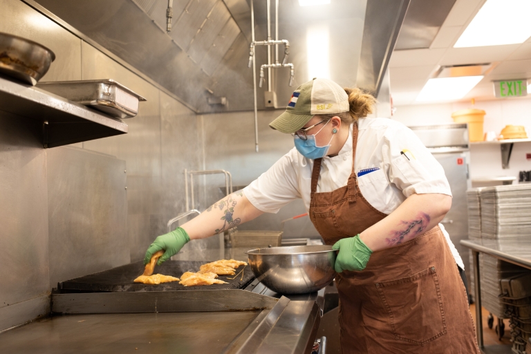 A female cook wearing a mask places chicken breasts on a grill in a kitchen.