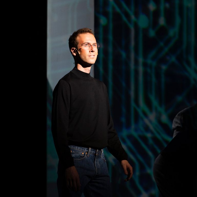 A man in a black turtleneck smirks on stage