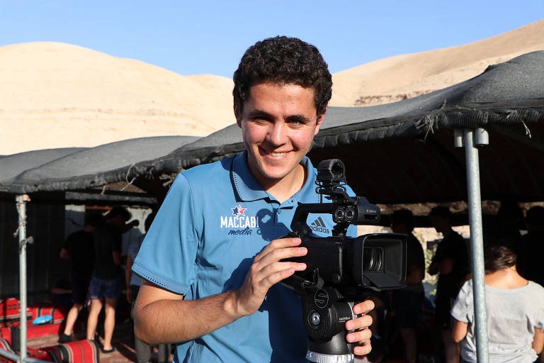 A young man stands behind a video camera smiling.