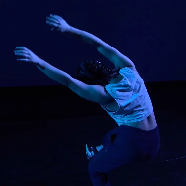 A dancer in blue light extends arms over her bowed head while jumping.