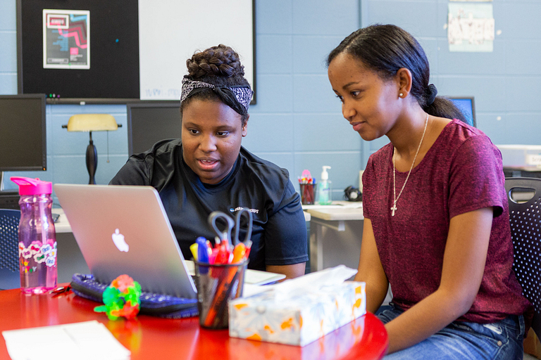 A student helps another with a paper. They are both looking at a laptop. 