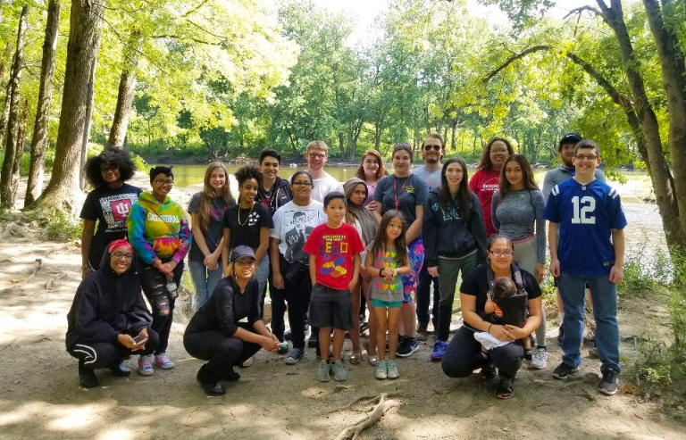 A large group of students from the Fostering Success Program stand by a wooded river.