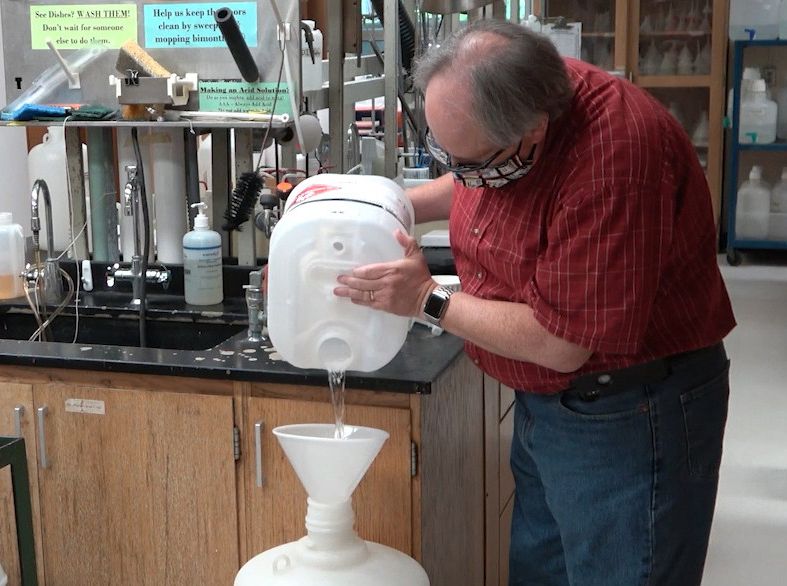 A man in a red plaid shirt pours chemicals for hand sanitizer into a large white container.
