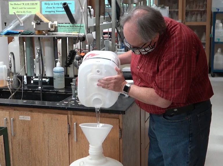 A man in a red plaid shirt pours chemicals for hand sanitizer into a large white container.