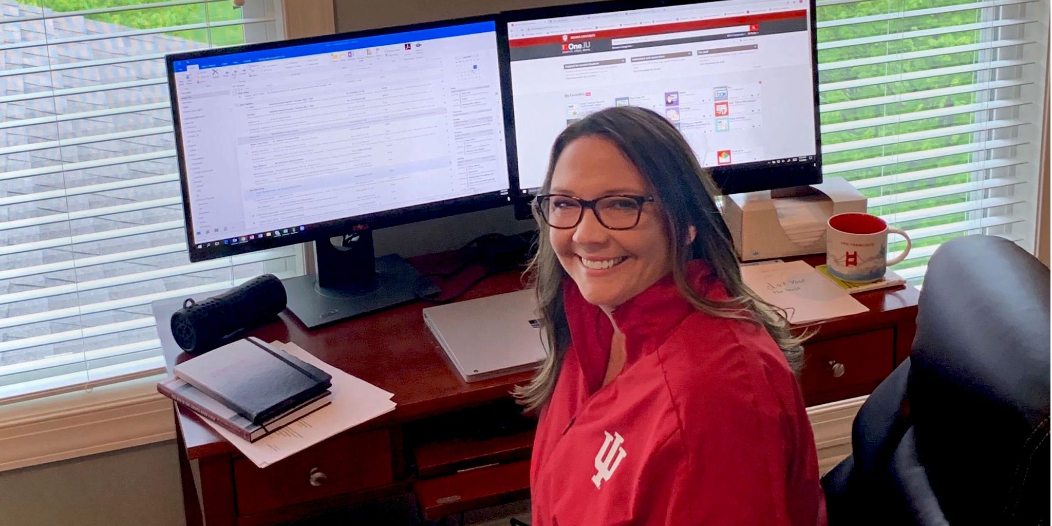 A woman wearing a red IU jacket sits at a computer desk in her home.