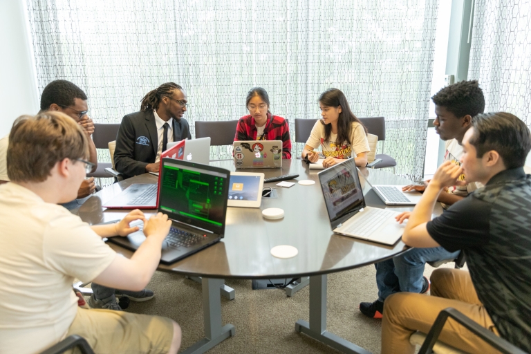 A group of seven students seated at a round table, studying with laptops