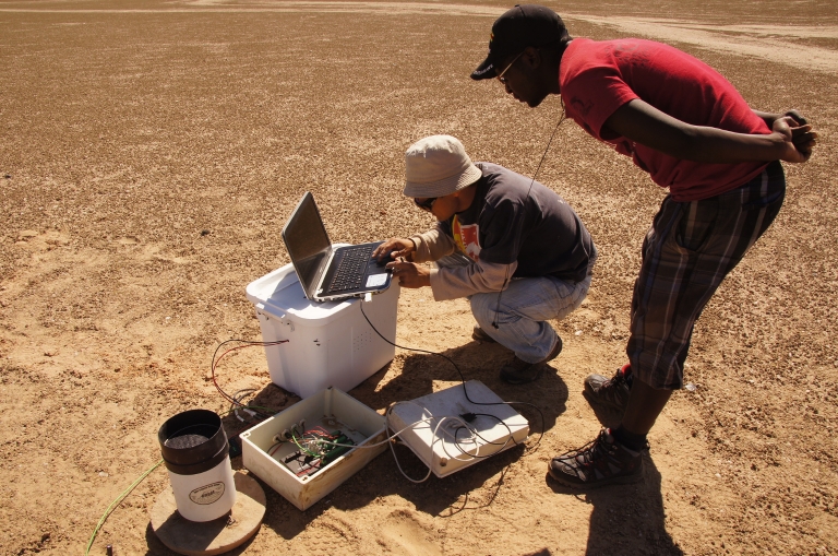 Lixin Wang kneels in Namib Desert to check a computer as a graduate student looks over his shoulder
